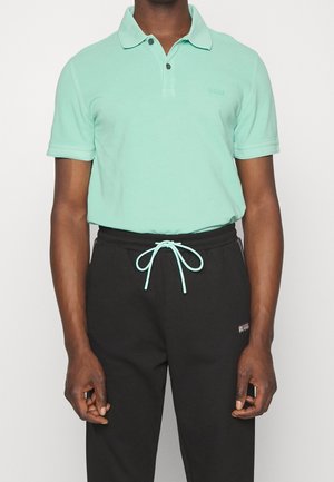 Mann trägt mintgrünes Poloshirt, das in schwarze Jogginghose mit Kordelzug, der in der Taille zur Schleife gebunden ist, gesteckt ist, vor weißem Hintergrund.