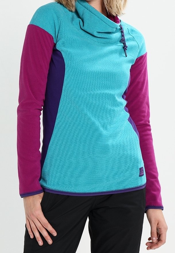 Femme portant un pullover à manches longues turquoise, violet et magenta avec bloc de couleurs, col bénitier à cordon et pantalon noir.