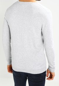 Homme portant un pull léger gris clair à manches longues et un jean bleu foncé, vu de dos sur un fond clair uni.
