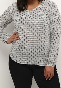 Femme portant un haut à manches longues blanc avec des motifs géométriques noirs et un pantalon noir, posant avec une main sur la hanche.