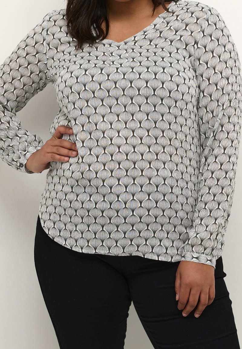 Femme portant un haut à manches longues blanc avec des motifs géométriques noirs et un pantalon noir, posant avec une main sur la hanche.