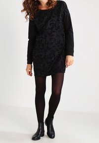 Schwarzes Tunika-Kleid mit strukturiertem floralen Stickmuster auf der Vorderseite, langen Ärmeln und kombiniert mit schwarzen Strumpfhosen und Ankle Boots.