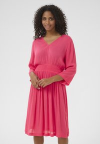 Robe rose corail avec un décolleté en V, des manches trois-quarts et une taille froncée. Tissu doux et léger avec une jupe fluide.