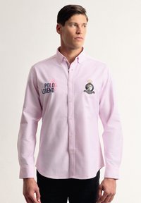 Camisa de botones de manga larga en rosa claro que presenta el texto bordado "Polo Legend" y un escudo en el área del pecho izquierdo. Tela de algodón suave.