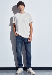 Joven de pie contra una pared blanca, vistiendo una camiseta blanca sencilla, jeans azules y zapatillas blancas, con una mano en el bolsillo.