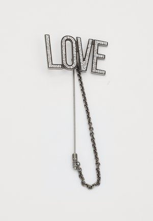 Kovový klopový odznak se slovem "LOVE" z průhledných rhinestones, opatřený stříbrným řetízkem na připevnění, na bílém pozadí.