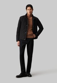 Chaqueta negra acolchada sobre un suéter marrón claro, combinada con pantalones oscuros y zapatos negros. La chaqueta tiene botones a presión y cuello.