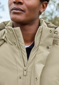 Beige Kapuzenjacke mit Reißverschluss und Druckknöpfen, aus strukturiertem Stoff, mit einer Nahaufnahme des Kragens und des Kinns.