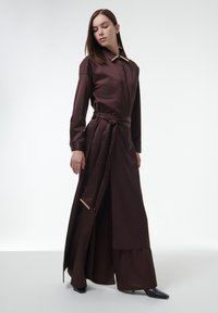 Lange bruine jurk met een kraaghalslijn, een strakke taille, en gouden accenten. De stof is glad met een gelaagd, vloeiend silhouette.