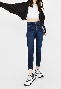 Jeans azul de cintura alta con cierre de cinco botones, combinados con un top corto blanco y una chaqueta bomber negra. Zapatillas completan el conjunto.