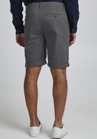 Gray cotton shorts with rolled cuffs, back pockets. Model wearing white sneakers, blue button-up shirt, with a neutral background.