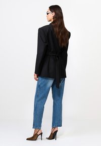 Naine pikkade pruunide juustega, kandes musta vöödiga bleiserit, siniseid teksaseid, leopardimustriga kõrgeid kontsi ja päikeseprille, seismas külg ees valge tausta ees.