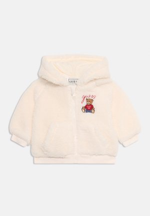 Hoodie za otroke v smetanasti barvi, puhasto, z zadrgo spredaj, kenguru žepi in vezene medvedek oblečen v rdečo majico s tekstom "guess" zgoraj.