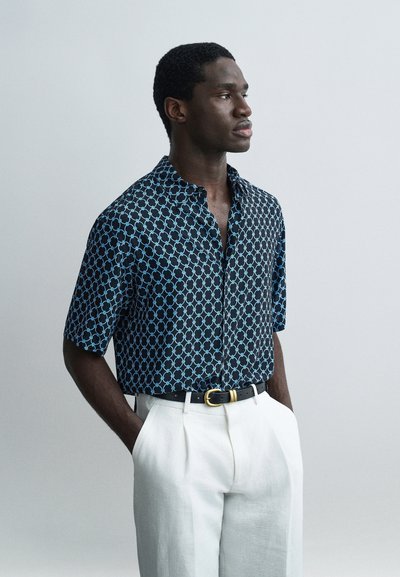 Homme aux cheveux courts et bouclés portant une chemise à manches courtes à motifs rentrée dans un pantalon blanc, les mains dans les poches, faisant face à droite.