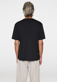 Man med grått hår som bär en lös svart kortärmad skjorta och ljusgrå byxor, stående vänd bortåt mot en enkel vit bakgrund.