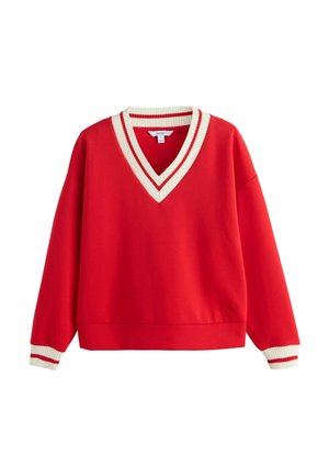 Kvinde med mørkt hår i en knold iført en rød sweater med hvide og røde stribede manchetter, en blå stribet skjorte under og et cremefarvet lagdelt skørt.