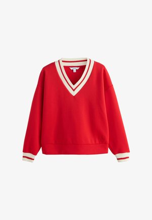 Donna con capelli scuri raccolti in uno chignon, indossa un maglione rosso con polsini a righe bianche e rosse, una camicia a righe blu sotto e una gonna crema a balze.