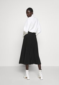 White, oversized sweater with long sleeves paired with a black, pleated midi skirt. Model wears white ankle boots with a block heel.