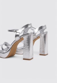 Sandales à talons hauts en argent avec une finition texturée, un large talon bloc et des lanières entrecroisées. Comprend une sangle de cheville réglable et une semelle beige.