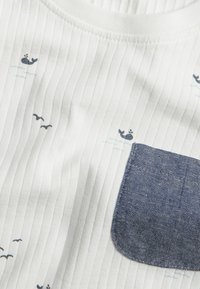 Weißes geripptes Shirt mit einer blauen Denim-Tasche, verziert mit kleinen Walen und Vogelmustern in Blau. Strukturierter Stoff mit kontrastierender Taschendetail.