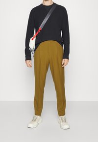 Homme portant une chemise noire à manches longues, un pantalon moutarde, des baskets blanches et un sac bandoulière blanc avec une sangle rouge et noire.