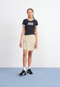 Tricou negru cu accente albe și logo-ul "PUMA", asociat cu o fustă scurtă bej. Îmbrăcat cu pantofi sport negri și șosete albe până la glezne.