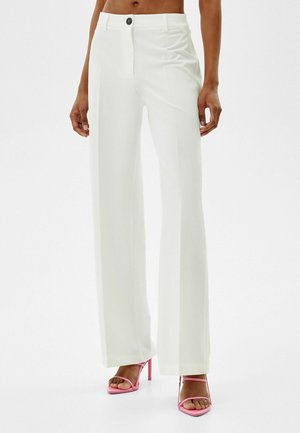 Vrouw die witte, op maat gemaakte wijd uitlopende broek en roze sandalen met hoge hak draagt, staand tegen een effen achtergrond.