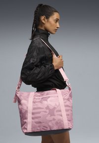 Sac fourre-tout rose à motif camouflage avec poignées doubles, bandoulière réglable et poche zippée. Fabriqué en tissu durable à la texture lisse.