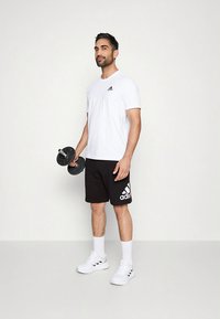 Maglietta sportiva bianca con logo nero, pantaloncini neri con dettagli bianchi, che tiene un manubrio nero, in piedi su una superficie chiara.