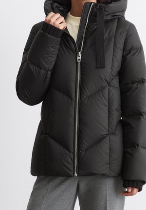 Personne portant une doudoune noire matelassée avec capuche et fermeture éclair argentée, associée à un pantalon gris et une chemise intérieure beige.