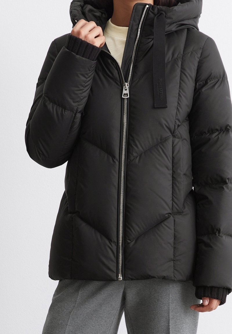 Personne portant une doudoune noire matelassée avec capuche et fermeture éclair argentée, associée à un pantalon gris et une chemise intérieure beige.