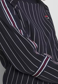 Sudadera navy con rayas verticales blancas, cuello de canalé y logotipo de marca. Presenta acentos en rayas rojas, blancas y azules en los puños.