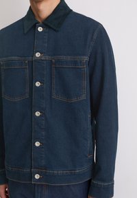 Donkerblauwe spijkerjack met een corduroy kraag, twee borstzakken, knoopsluiting aan de voorkant en contrasterende oranje stiksels.