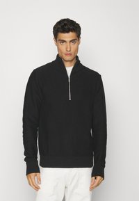 Hanorac negru cu fermoar, cu guler și manșete cu aspect texturat, dotat cu un fermoar de lungime medie și design din tricot. Purtați peste un tricou alb.