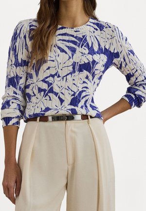 Kobieta w niebiesko-białym swetrze w kwiatowy wzór z długim rękawem, wpuszczonym w wysokowstanowe beżowe spodnie z brązowym paskiem.