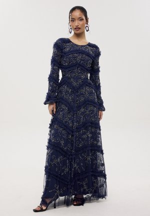 Mujer con un vestido largo azul marino con bordado floral y franjas horizontales con volantes, combinado con tacones abiertos negros y pendientes coloridos.