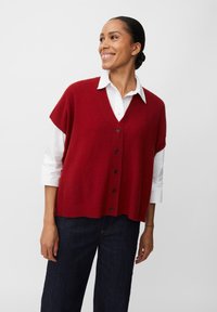 Gilet rosso a maglia con scollo a V e chiusura con bottoni, indossato sopra una camicia bianca con colletto. Texture morbida e vestibilità rilassata, abbinato a jeans scuri.