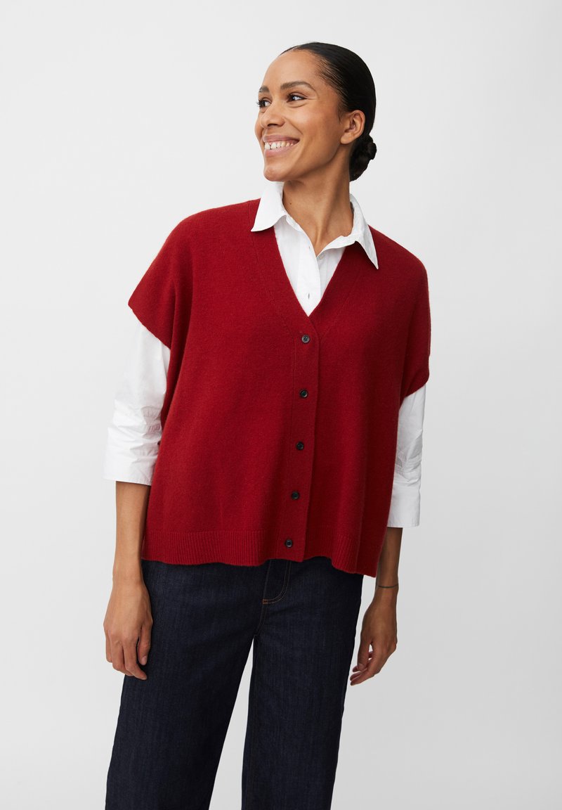 Gilet rosso a maglia con scollo a V e chiusura con bottoni, indossato sopra una camicia bianca con colletto. Texture morbida e vestibilità rilassata, abbinato a jeans scuri.