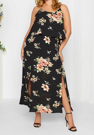 Femme portant une robe longue noire sans manches à motifs floraux, avec des fentes latérales et un haut superposé, associée à des sandales à talons hauts noires.