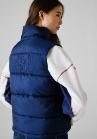 Ναυτικό γιλέκο με υψηλό γιακά και ανάγλυφο λογότυπο "Red Bull RACING" στην πλάτη. Διαθέτει μανίκια με λευκές και κόκκινες λεπτομέρειες και λεία υφή.