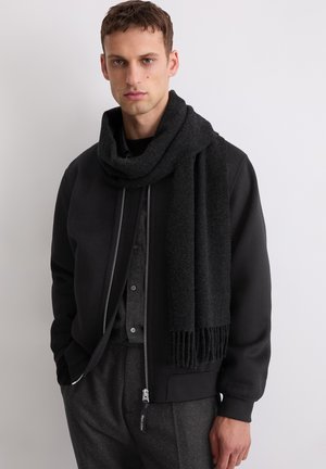 Écharpe en laine noire avec des extrémités à franges, drapée sur une veste bomber noire et une chemise sombre, avec une fermeture éclair à l'avant et un tissu texturé.