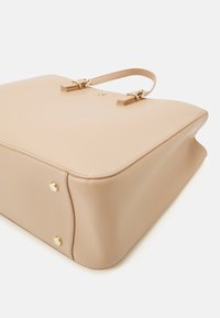 Sac à main en cuir beige avec une finition texturée, quincaillerie dorée et double anses supérieures. Présente une silhouette structurée et des pieds en métal protecteurs.