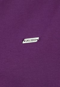 Un tessuto viola con una texture a righe presenta un piccolo etichetta d'argento con scritto "SMILODOX" attaccata con filo viola.