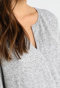 Femme avec de longs cheveux bruns portant un pull en tricot gris clair avec une subtile découpe en V.