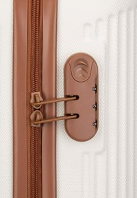 Hartschalenkoffer in Weiß mit strukturiertem Oberflächenfinish. Ausgestattet mit einem braunen Reißverschluss und einer Kombinationsschnalle, sowie zwei Reißverschlussschiebern für einen einfachen Zugriff.
