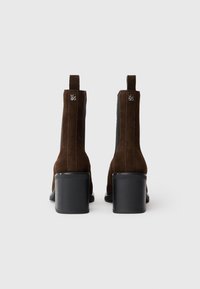 Botines de ante marrón con un tacón negro grueso, laterales elásticos interiores y tiradores traseros para ponérselos fácilmente. Textura suave y diseño aerodinámico.