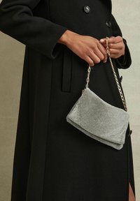 Borsa a pochette argentata con texture, di forma rettangolare, tenuta da una tracolla in catena dorata, indossata con un cappotto nero con bottoni grandi.