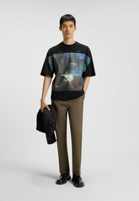 Een jonge man staat gekleed in een zwart oversized T-shirt met een grafische print, olijfgroene broeken, zwarte schoenen en houdt in één hand een zwarte jas.