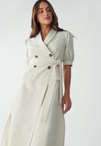 Vestido envolvente color crema con mangas cortas abullonadas, botones de doble botonadura, abertura lateral y cinturilla atada. Fabricado en una suave tela texturizada.