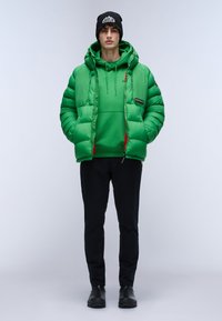 Chaqueta acolchada verde con capucha y acentos rojos, llevada sobre un hoodie verde a juego. Pantalones negros y zapatos negros completan el atuendo.
